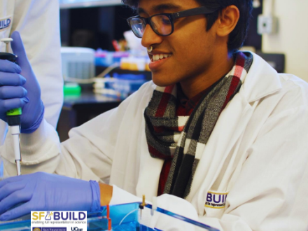 An SF BUILD scholar in a white lab coat using a pipette to put substance in a petri dish.